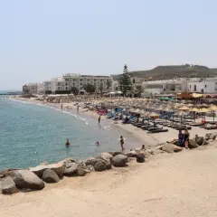 Słoneczna plaża na wyspie Kos z białymi hotelami, parasolami i turystami kąpiącymi się w morzu.
