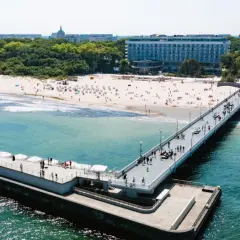 Molo i plaża w słoneczny dzień. W tle widać budynek sanatorium nad morzem, otoczony zielenią.