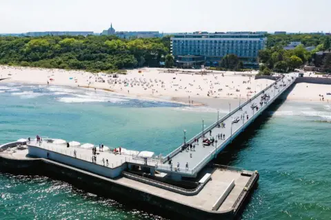 Molo i plaża w słoneczny dzień. W tle widać budynek sanatorium nad morzem, otoczony zielenią.