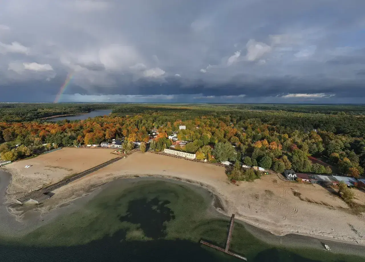 Jesienny krajobraz nad Skorzęcinem. Tęczowy łuk nad jeziorem i plażą, otoczone kolorowymi lasami.