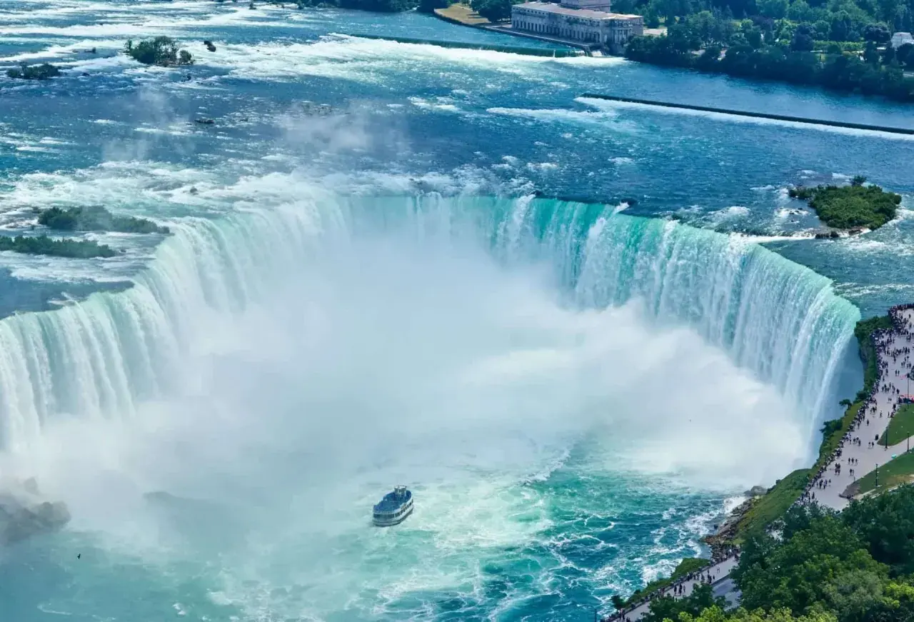 Potężny wodospad Niagara w całej okazałości. Widać łódź turystyczną i tłumy ludzi podziwiających ten cud natury.