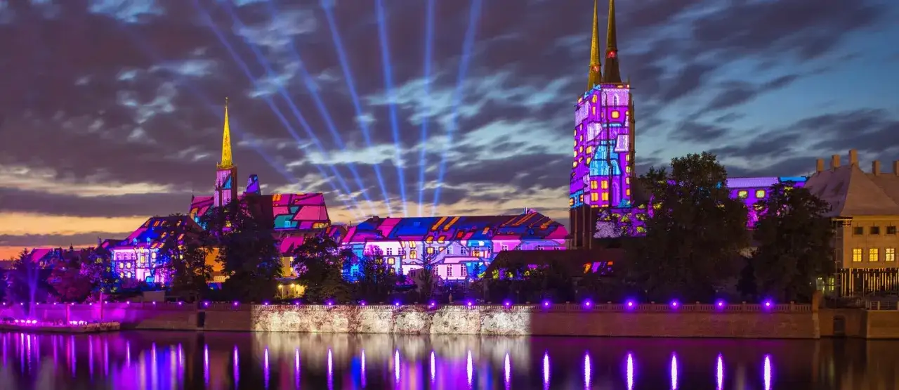 Wrocław nocą, niczym wenecja północy, rozświetlony feerią barw. Budynki i wieże katedry mienią się kolorowymi projekcjami, odbijając się w spokojnej tafli rzeki.