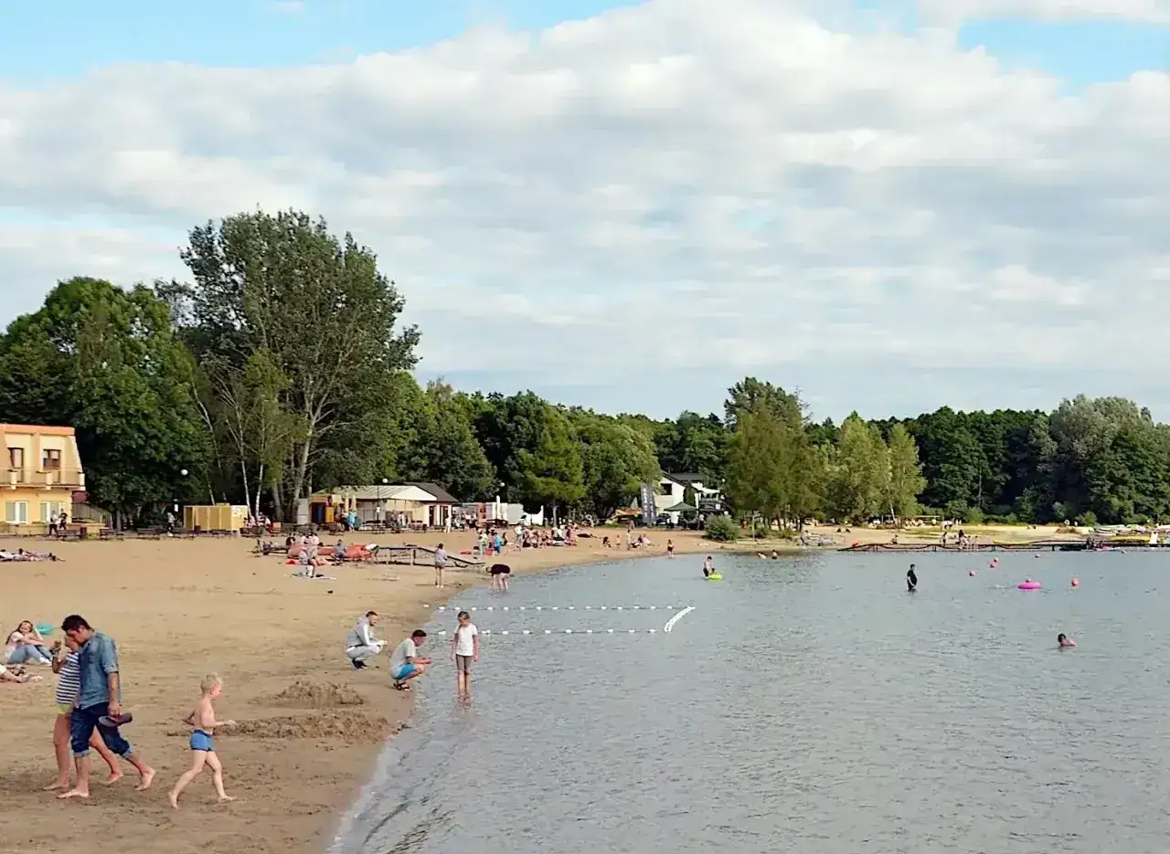 Słoneczny dzień nad jeziorem Skorzęcin. Ludzie odpoczywają na plaży, kąpią się i spacerują.