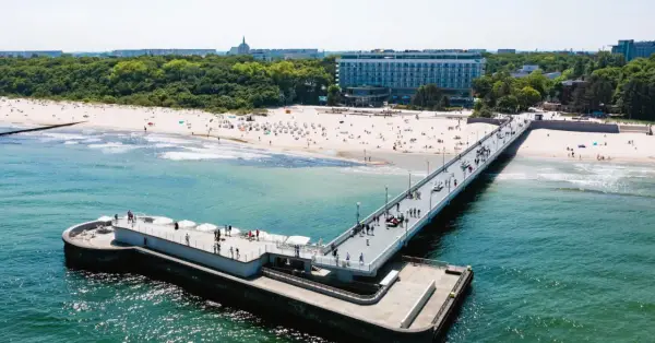 Molo i plaża w słoneczny dzień. W tle widać budynek sanatorium nad morzem, otoczony zielenią.