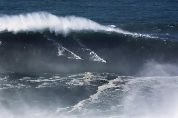 Surferzy ścigają się po gigantycznych nazare falach, tworząc białe smugi na ciemnym oceanie.
